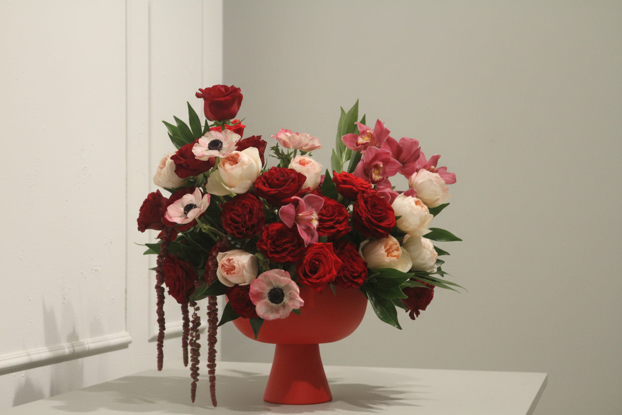 Arrangement of red, pink, and blush garden roses and orchids in a red compote against a white wall. 